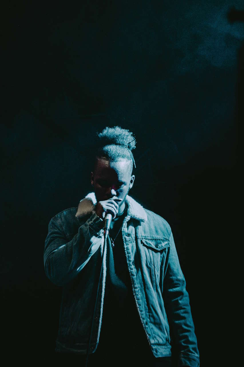 Ein Mann mit Afro-Zopf trägt eine Jeansjacke mit Fellkragen und hält ein Mikrofon vor einem dunklen Hintergrund.
