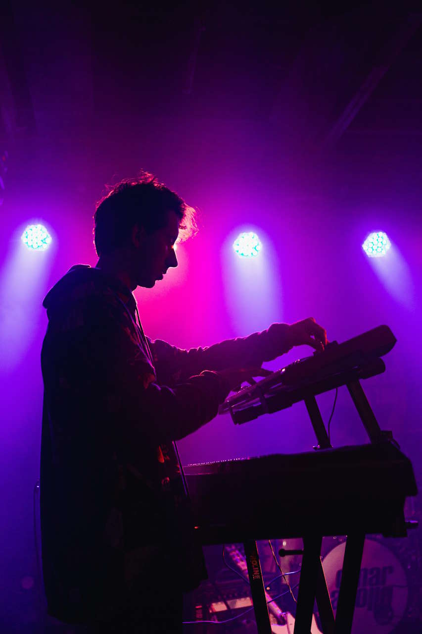 Silhouette eines Musikers, der an einem Keyboard spielt, beleuchtet von lila und pinken Bühnenlichtern mit drei hellen Spots im Hintergrund.