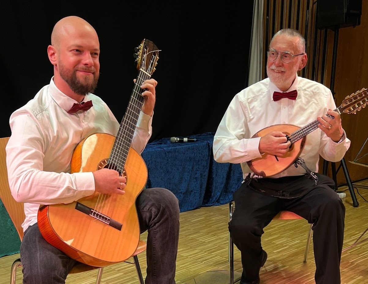 Zwei Männer in weißem Hemd mit roten Fliegen sitzen auf Stühlen und spielen Saiteninstrumente, links eine akustische Gitarre, rechts eine Mandoline, vor schwarzem Hintergrund und Holzboden.