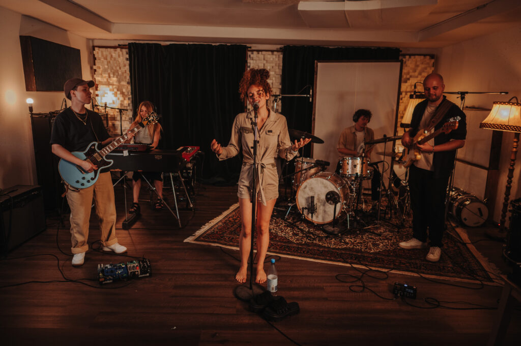 Fünfköpfige Band in einem Aufnahmeraum mit Holzfußboden und Teppich, besteht aus Sängerin in beigem Overall, Gitarrist mit blauer Gitarre, Keyboarderin, Schlagzeuger und Bassist, Instrumente und Mikrofone sind aufgebaut, gemütliche Beleuchtung durch Lampen und Studiopaneele im Hintergrund.