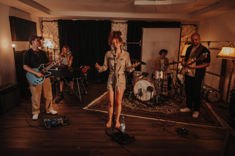 Fünfköpfige Band in einem Aufnahmeraum mit Holzfußboden und Teppich, besteht aus Sängerin in beigem Overall, Gitarrist mit blauer Gitarre, Keyboarderin, Schlagzeuger und Bassist, Instrumente und Mikrofone sind aufgebaut, gemütliche Beleuchtung durch Lampen und Studiopaneele im Hintergrund.
