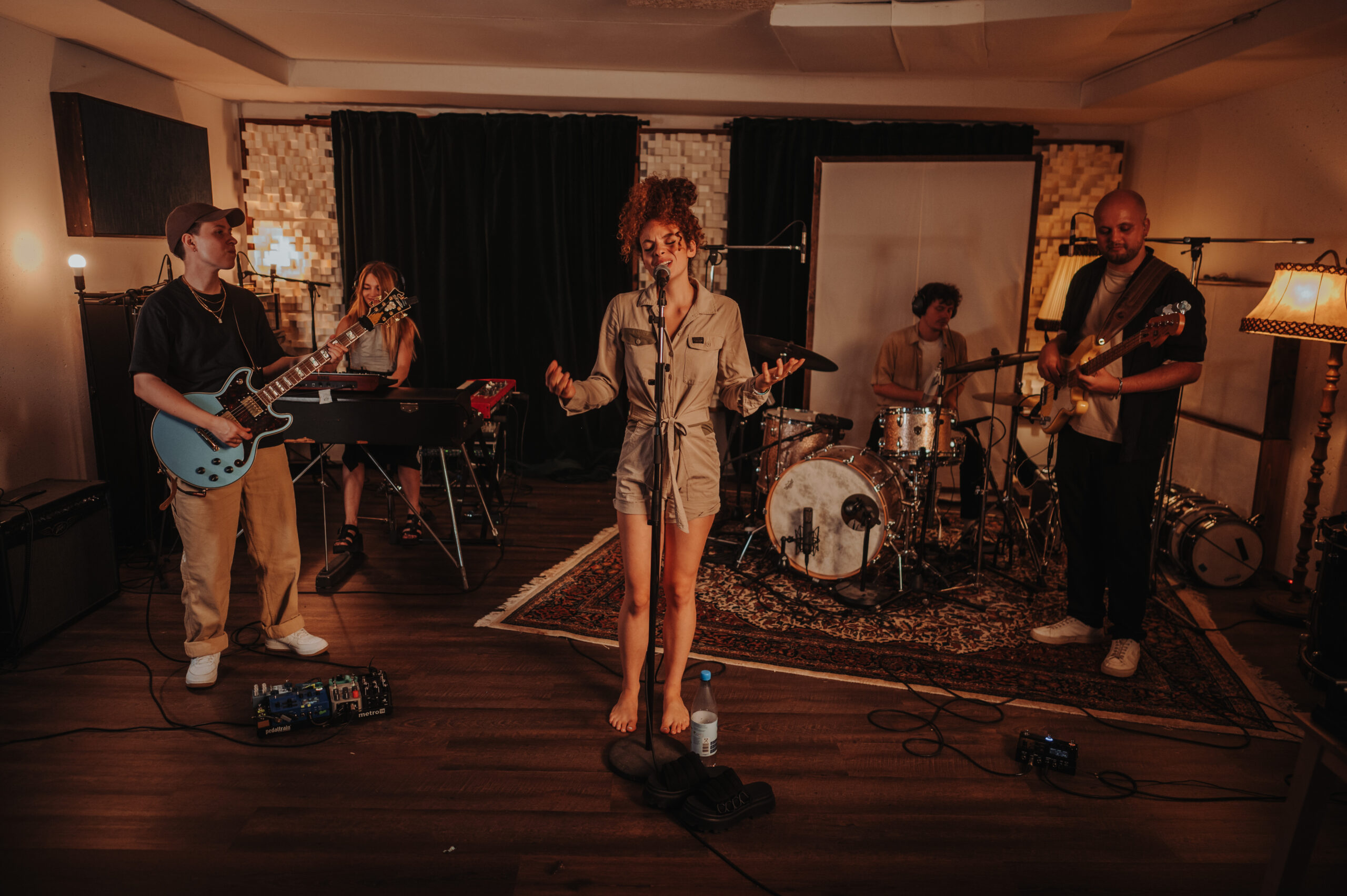 Fünfköpfige Band in einem Aufnahmeraum mit Holzfußboden und Teppich, besteht aus Sängerin in beigem Overall, Gitarrist mit blauer Gitarre, Keyboarderin, Schlagzeuger und Bassist, Instrumente und Mikrofone sind aufgebaut, gemütliche Beleuchtung durch Lampen und Studiopaneele im Hintergrund.