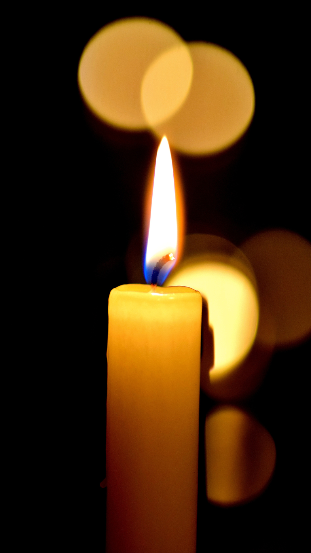 Brennende kerze mit Flamme vor einem schwarzen Hintergrund und verschwommenen hellgelben Lichtkreisen
