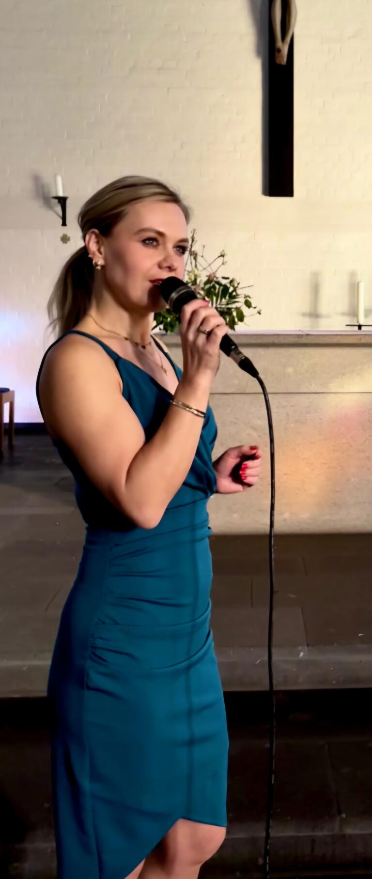 Frau mit blonden Haaren im blauen Kleid, die in einem Kirchenraum mit Mikrofon singt, vor einem Altar mit Blumen, Kerzen und einer Kreuzigungsdarstellung an der Wand.