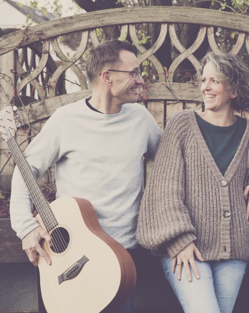 Mann mit Brille und Pullover hält eine Akustikgitarre und schaut lächelnd zu einer Frau mit lockigem Haar und dicker Strickjacke, die ebenfalls lächelt, beide lehnen an einem Holzzaun mit Ranken.