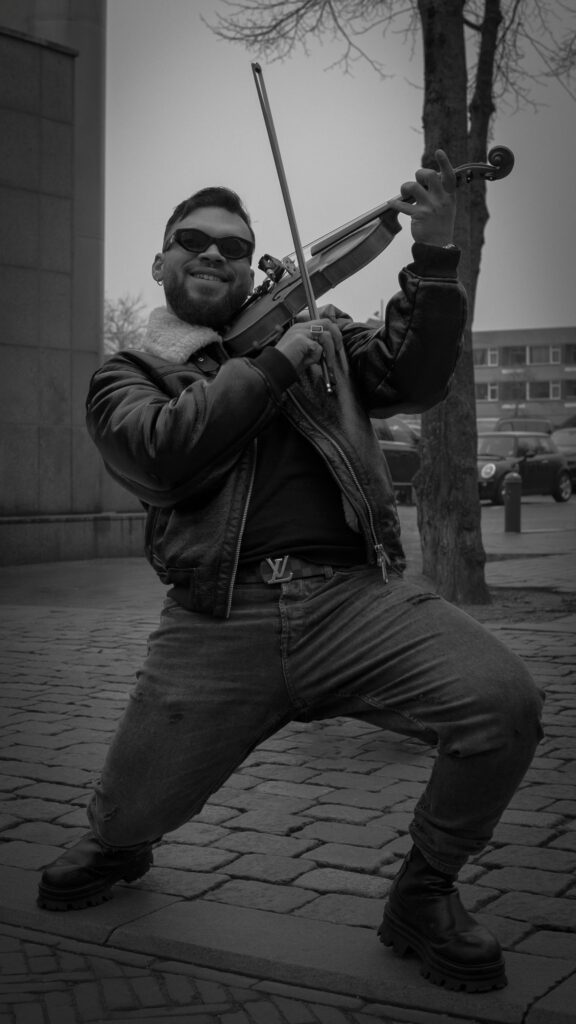 Ein Mann mit Sonnenbrille, Lederjacke und Stiefeln spielt auf der Straße energisch Geige und steht in einer weiten Pose auf Kopfsteinpflaster vor einer Baumreihe und parkenden Autos.