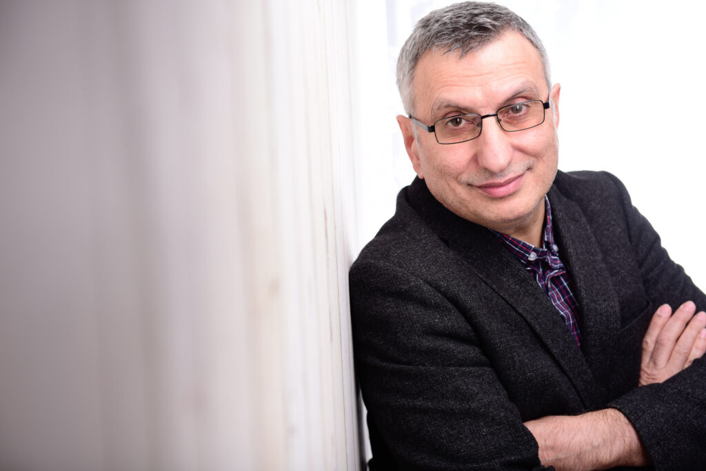 Mittelalter Mann mit kurzen grauen Haaren, Brille und schwarzem Sakko lehnt mit verschränkten Armen an eine helle Wand und lächelt in die Kamera.