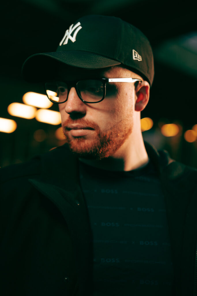 Porträt eines Mannes mit schwarzer New York Yankees Baseballkappe, braunem Bart, schwarzer Brille und dunkler Jacke, vor verschwommenem Hintergrund mit warmen Lichtern.