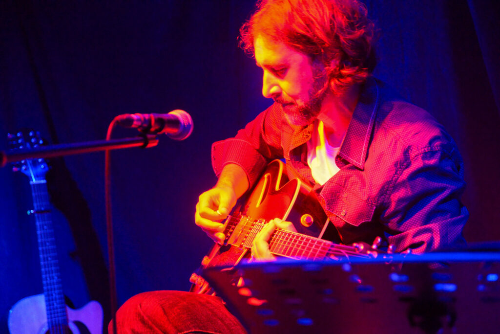 Mann mit Bart und langen Haaren spielt E-Gitarre auf einer Bühne mit bunter Bühnenbeleuchtung in Rot und Blau, daneben ein Mikrofon und im Hintergrund eine Akustikgitarre.