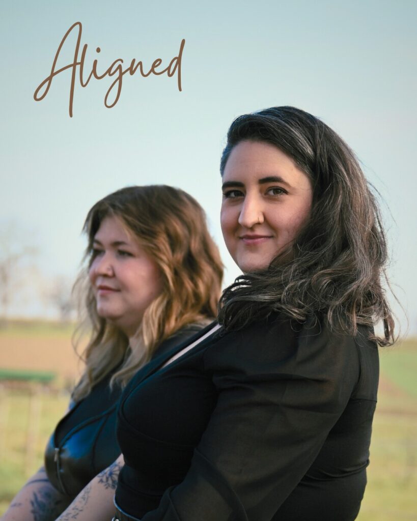 Zwei Frauen vor unscharfem Hintergrund mit Landschaft, die rechte Frau mit dunklem Haar blickt in die Kamera, die linke mit hellbraunem Haar sieht seitlich weg, darüber der Schriftzug 'Aligned' in handschriftlicher Schrift.