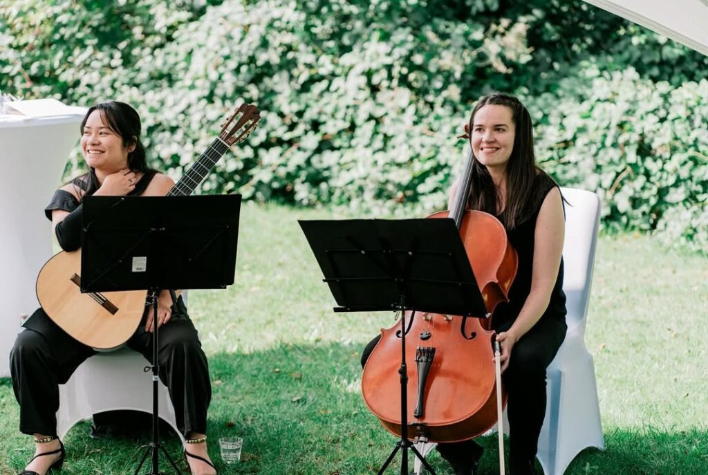Zwei Musikerinnen sitzen draußen auf weißen Stühlen auf einem grünen Rasen, eine hält eine Gitarre, die andere ein Cello, beide lächeln und haben Notenständer vor sich.