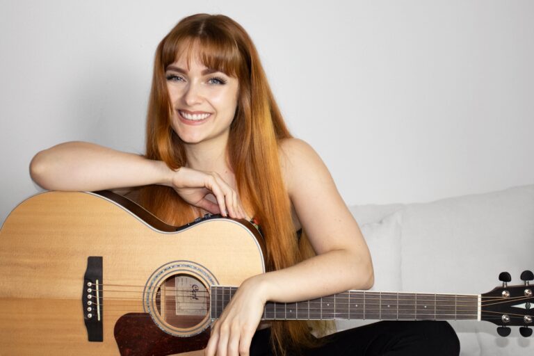 Frau mit roten langen Haaren, die lächelnd mit einer Akustikgitarre auf einem hellen Sofa sitzt und die Gitarre umarmt.