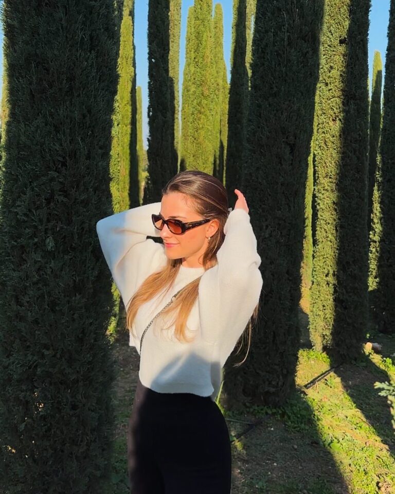 Junge Frau mit braunen langen Haaren und Sonnenbrille, die auf einem sonnenbeschienenen Weg zwischen hohen, dünnen Zypressenbäumen steht und die Hände hinter den Kopf nimmt, trägt einen weißen Pullover und schwarze Hose.