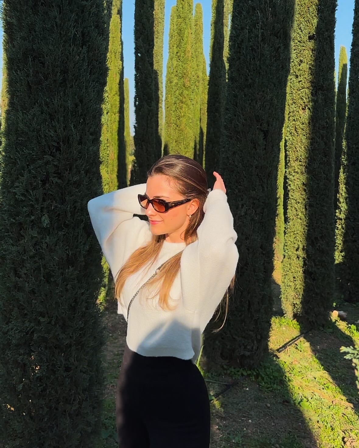 Junge Frau mit braunen langen Haaren und Sonnenbrille, die auf einem sonnenbeschienenen Weg zwischen hohen, dünnen Zypressenbäumen steht und die Hände hinter den Kopf nimmt, trägt einen weißen Pullover und schwarze Hose.