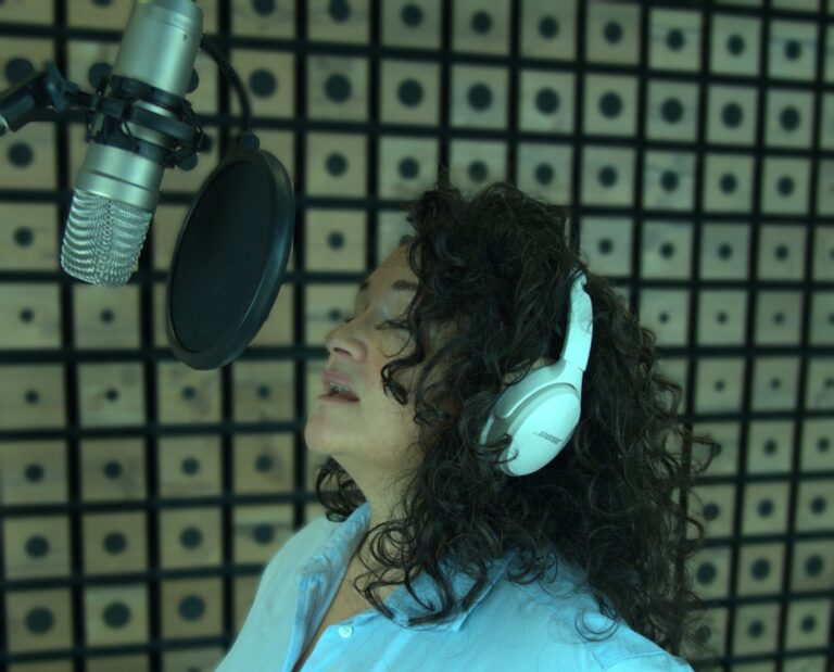 Frau mit langen lockigen dunklen Haaren und weißen BOSE-Kopfhörern, die in einem Aufnahmestudio in ein Mikrofon singt, vor einer Wand mit schallabsorbierenden Paneelen.