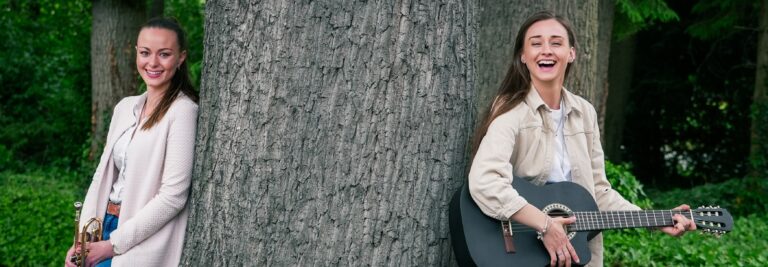 Zwei junge Frauen lehnen an einem großen Baum, die eine hält eine Trompete, die andere spielt eine schwarze Gitarre, beide lachen und befinden sich in einem grünen Park.