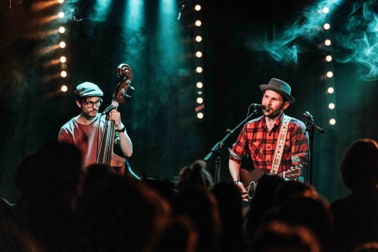 Zwei Musiker auf einer Bühne bei einem Konzert, links ein Kontrabassist mit Brille und Mütze, rechts ein Sänger mit Gitarre und Hut, umgeben von blaugrüner Bühnenbeleuchtung und Publikum im Vordergrund