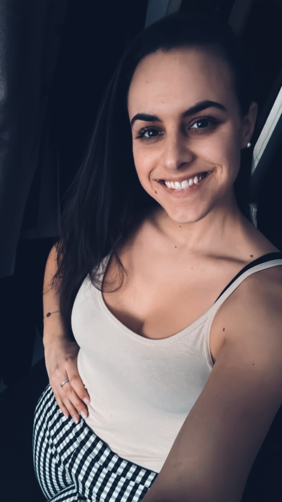 Lächende Frau mit langen dunklen Haaren, die eine weiße ärmellose Bluse und schwarz-weiß karierte Hose trägt, mit der rechten Hand auf ihrem Bauch