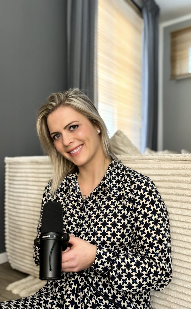 Frau mit blonden Haaren und geblümtem Hemd sitzt auf einem hellen Sofa und hält ein schwarzes Mikrofon in der Hand, im Hintergrund sind Fenster mit Jalousien und graue Vorhänge zu sehen