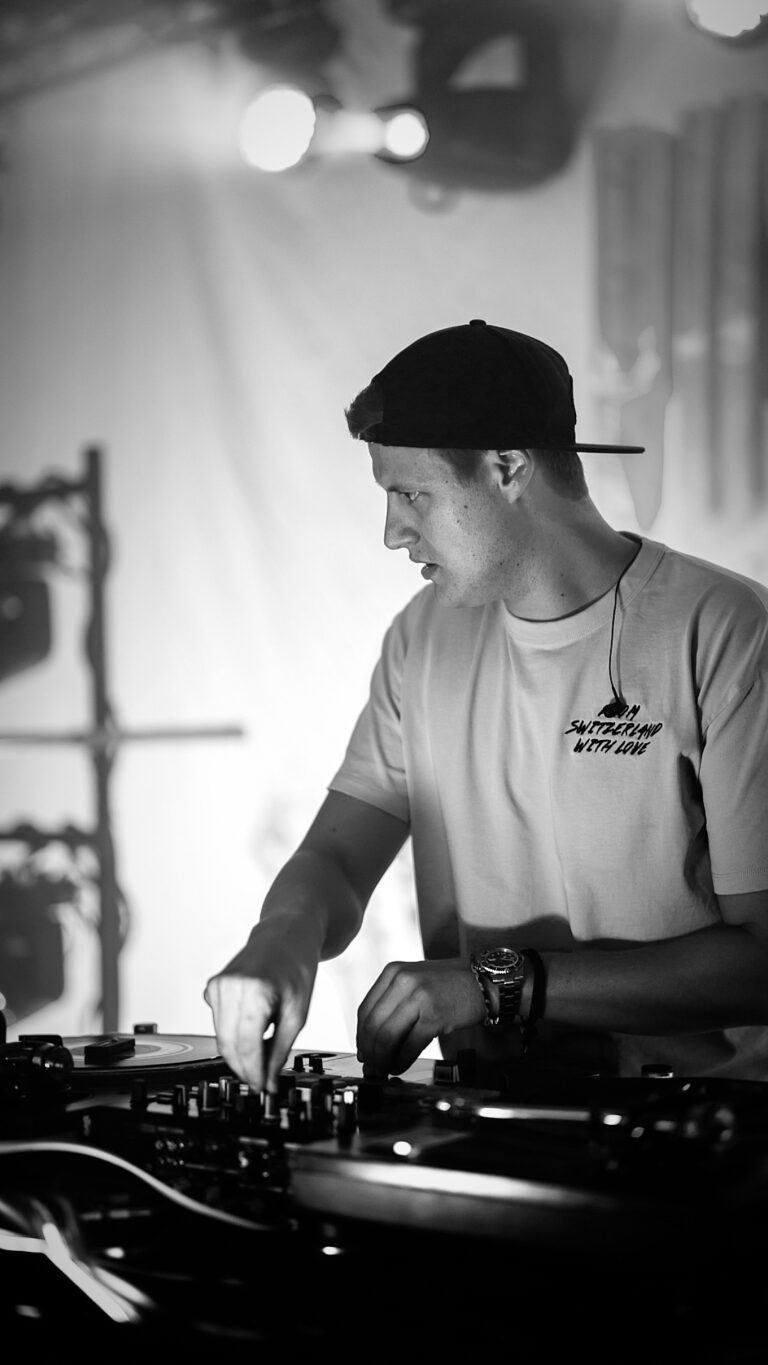 Schwarz-weiß-Foto eines jungen DJs mit rückwärts getragenem Baseballcap, der konzentriert an seinem Mischpult arbeitet und Knöpfe einstellt, im Hintergrund Bühnenlicht und verschwommene Ausrüstung.