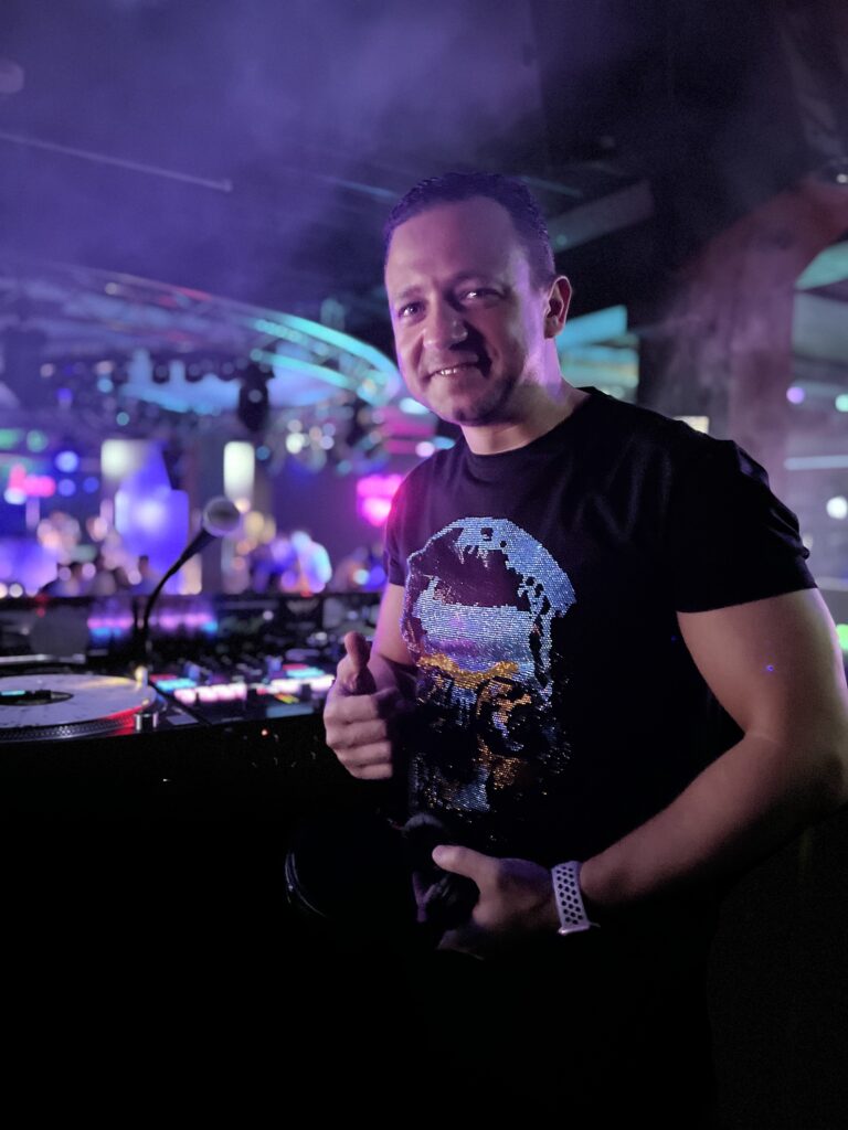 Mann in schwarzem T-Shirt mit Totenkopf-Motiv steht lächelnd vor einem DJ-Pult in einem Club mit bunter Bühnenbeleuchtung und rauchiger Atmosphäre.