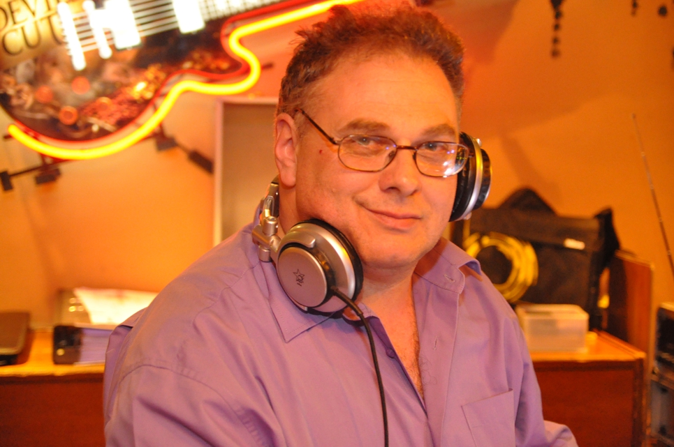 Mann mit Brille und Kopfhörern um den Hals trägt ein hellviolettes Hemd, im Hintergrund sind verschwommene Gegenstände und eine bunte Leuchtreklame zu sehen.