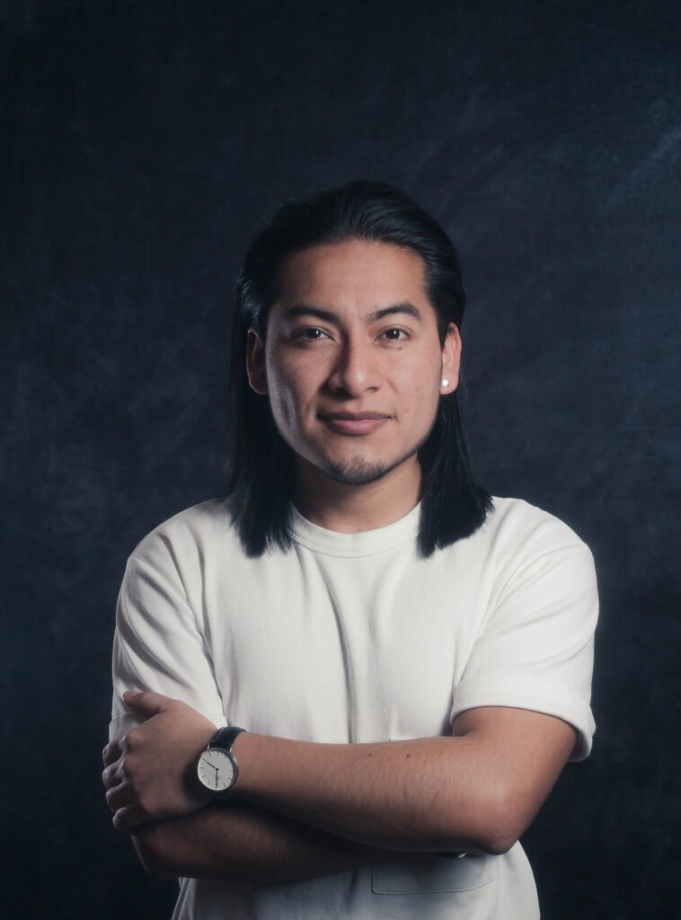 Junger Mann mit langen schwarzen Haaren, weißem T-Shirt und Armbanduhr, der die Arme verschränkt vor dunklem Hintergrund schaut.