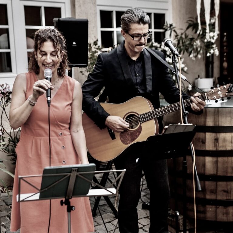 Eine Frau im ärmellosen roten Kleid singt in ein Mikrofon, neben ihr spielt ein Mann mit Brille und Schnurrbart eine akustische Gitarre, beide stehen vor einem Fenstermauerwerk mit Pflanzen und einer Holztischdekoration im Hintergrund.