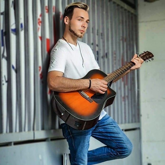 Ein Mann mit blond-schwarzem Haar und Bart trägt ein weißes T-Shirt und blaue Jeans und spielt eine braune Akustikgitarre vor einer Wand mit metallischen vertikalen Rohren.