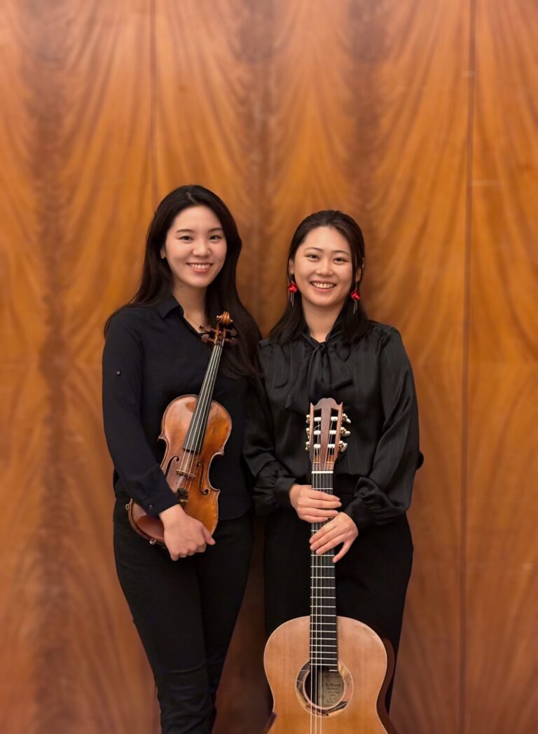 Zwei junge Frauen in schwarzen Outfits stehen vor einer Holzwand, die linke hält eine Geige, die rechte eine Gitarre, beide lächeln in die Kamera.