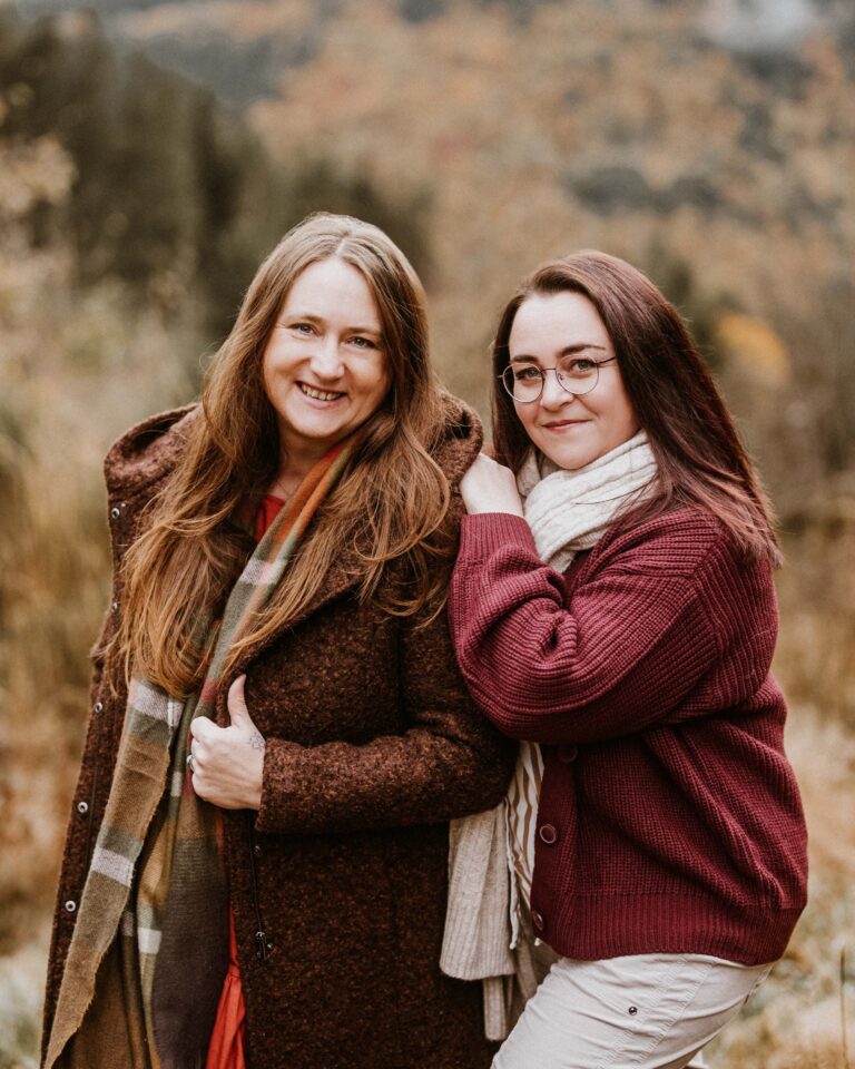 Zwei Frauen stehen nah beieinander im Freien, beide lächeln; eine trägt einen braunen Mantel und Schal, die andere eine weinrote Strickjacke, Schal und Brille, herbstliche Waldlandschaft im Hintergrund.