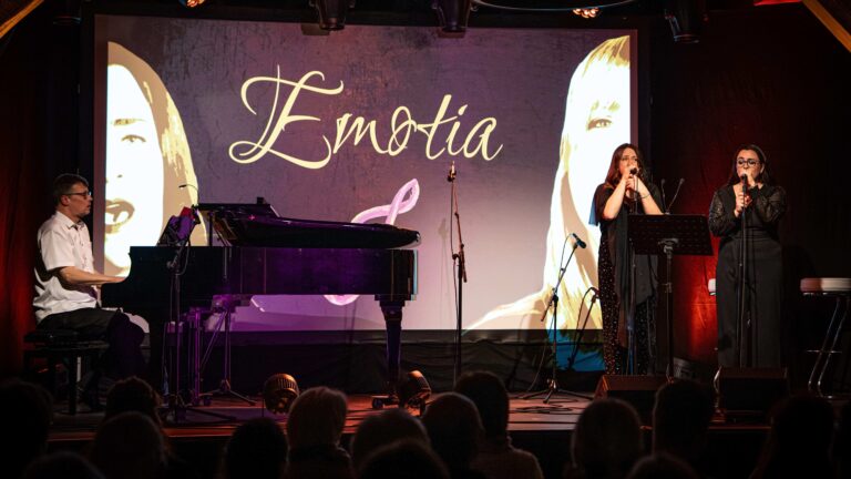 Musikperformance auf einer Bühne mit einem Pianisten links, zwei Sängerinnen rechts vor einem großen Bildschirm mit dem Schriftzug "Emotia" und Porträts von Frauen im Hintergrund, Zuschauer im Vordergrund.