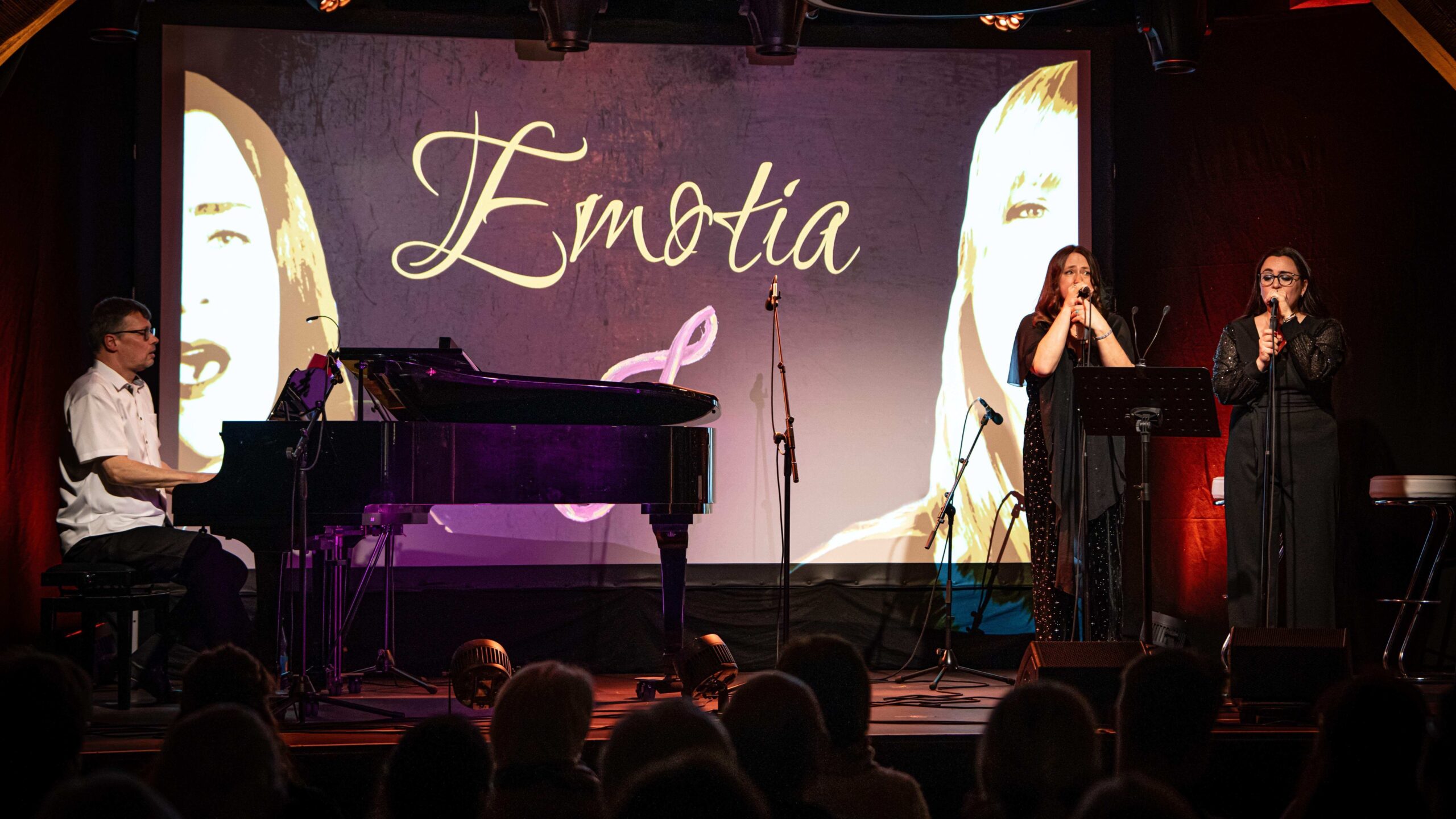 Musikperformance auf einer Bühne mit einem Pianisten links, zwei Sängerinnen rechts vor einem großen Bildschirm mit dem Schriftzug "Emotia" und Porträts von Frauen im Hintergrund, Zuschauer im Vordergrund.