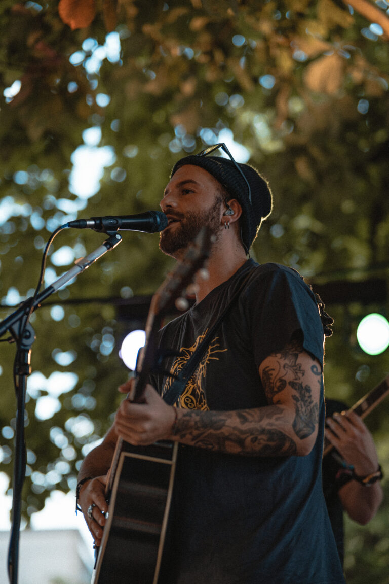 Mann mit schwarzen Tattoos an beiden Armen spielt Gitarre und singt in ein Mikrofon, trägt Beanie, dunkles T-Shirt mit gelbem Aufdruck und Ohrhörer, im Hintergrund verschwommene grüne Blätter und Lichtpunkte.