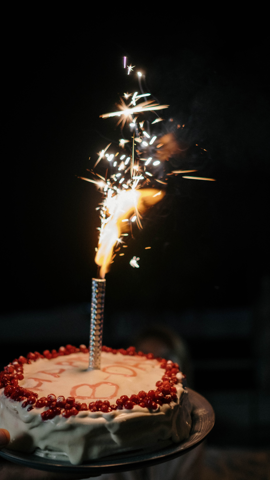 Geburtstagstorte mit weißem Zuckerguss und roter Beerenverzierung, darauf eine brennende Wunderkerze vor dunklem Hintergrund