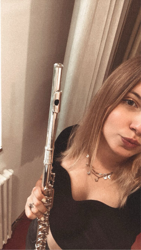 Junge Frau mit mittellangen blonden Haaren, die eine silberne Querflöte hält, trägt ein schwarzes Oberteil und eine Halskette mit Schmetterlingsanhängern, aufgenommen vor einem Hintergrund mit Gardinen und einem Heizkörper.