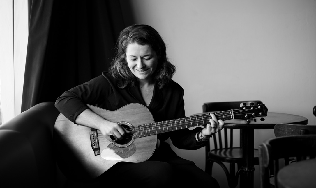 Schwarz-weiß-Foto einer lächelnden Frau, die auf einem Stuhl sitzt und eine akustische Gitarre spielt, mit einem dunklen Vorhang und mehreren Tischen und Stühlen im Hintergrund.