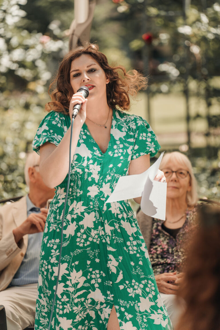 Frau mit braunen Locken und grünem, weiß geblümtem Kleid hält ein Mikrofon und ein Blatt Papier, im Hintergrund sitzen zwei ältere Personen in einem Garten oder Park