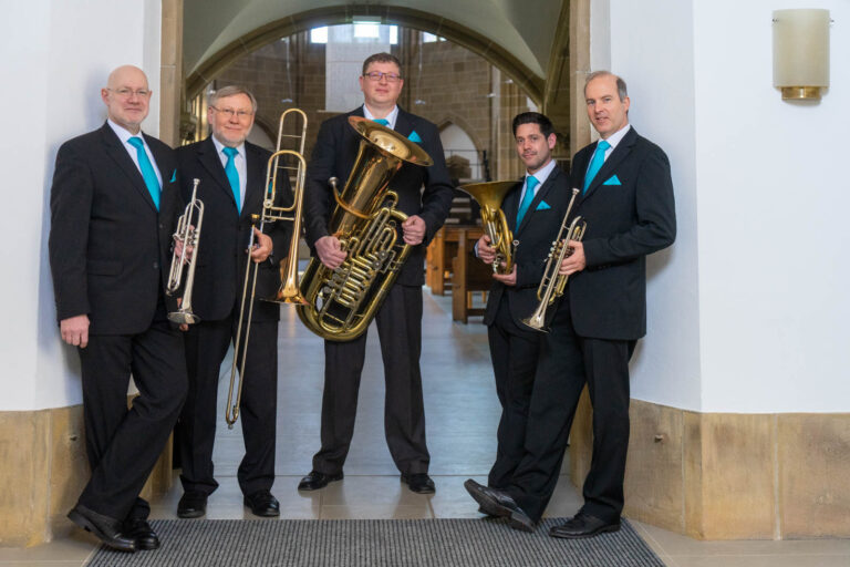 Fünf Männer in schwarzen Anzügen mit hellblauen Krawatten und Einstecktüchern, die in einem Kircheninneren mit Bogen gewölbter Decke und Sandsteinwänden stehen, jeweils mit Blechblasinstrumenten (Trompete, Posaune, Tuba und Horn) in der Hand.