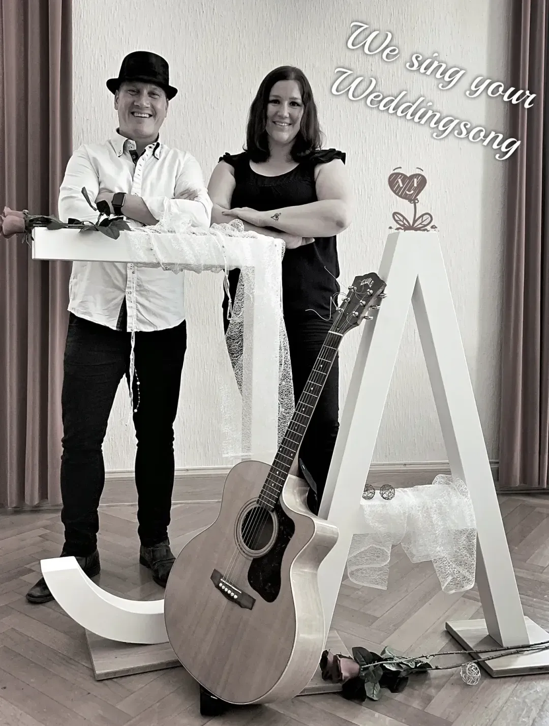 Ein Mann mit schwarzem Hut und weißem Hemd sowie eine Frau in schwarzem Oberteil stehen hinter großen weißen Buchstaben J und A, eine Gitarre lehnt an den Buchstaben, eine rosa Rose liegt am Boden, im Hintergrund Text We sing your Weddingsong.