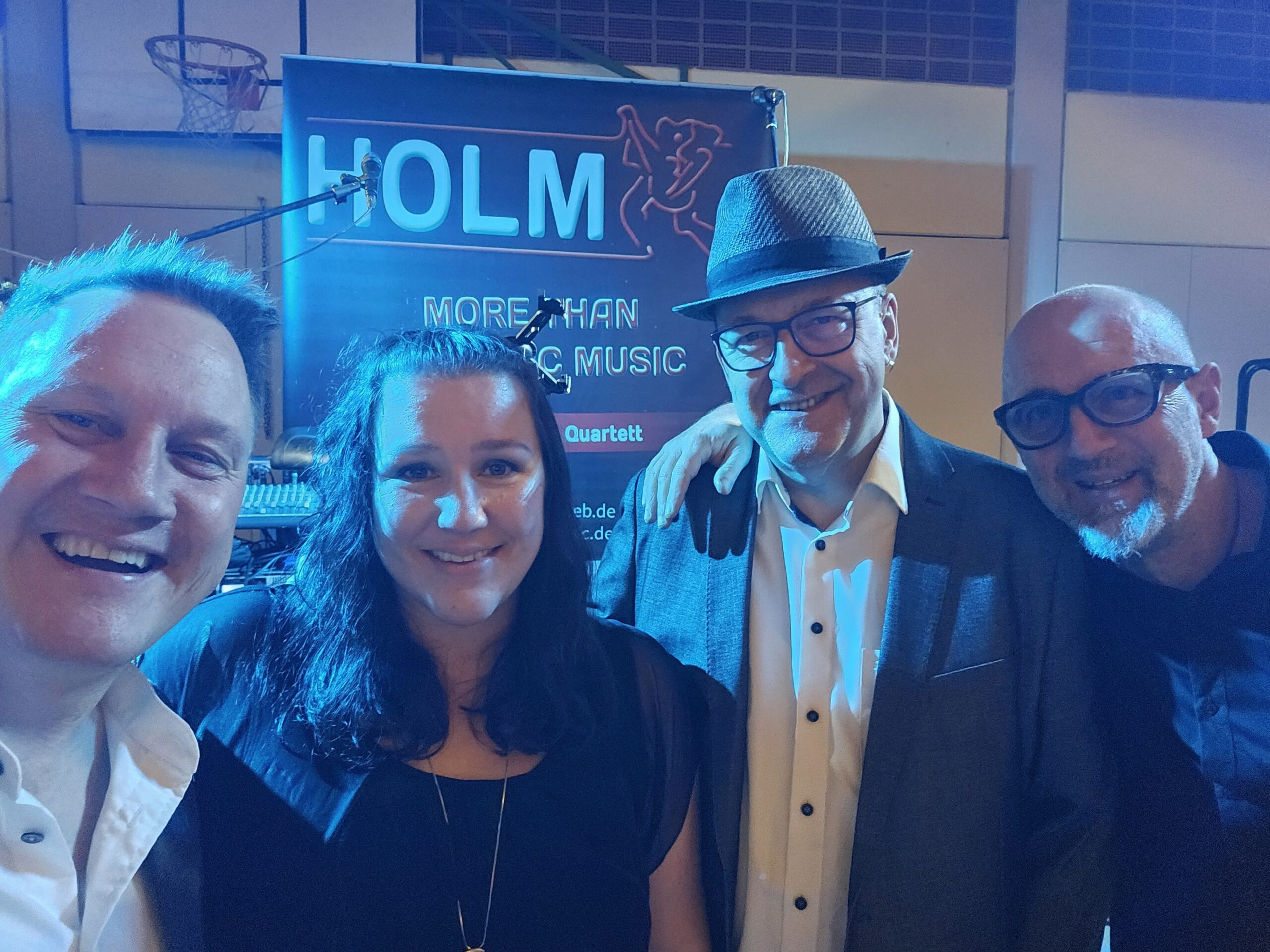 Vier lächelnde Personen vor einem blauen Werbebanner mit der Aufschrift HOLM und mehr Musik, darunter ein Mann mit grauem Hut und Brille, eine Frau mit schwarzem Oberteil und Halskette sowie zwei Männer mit hellen Hemden und dunklen Brillen in einer Innenraumhalle mit Basketballkorb im Hintergrund.