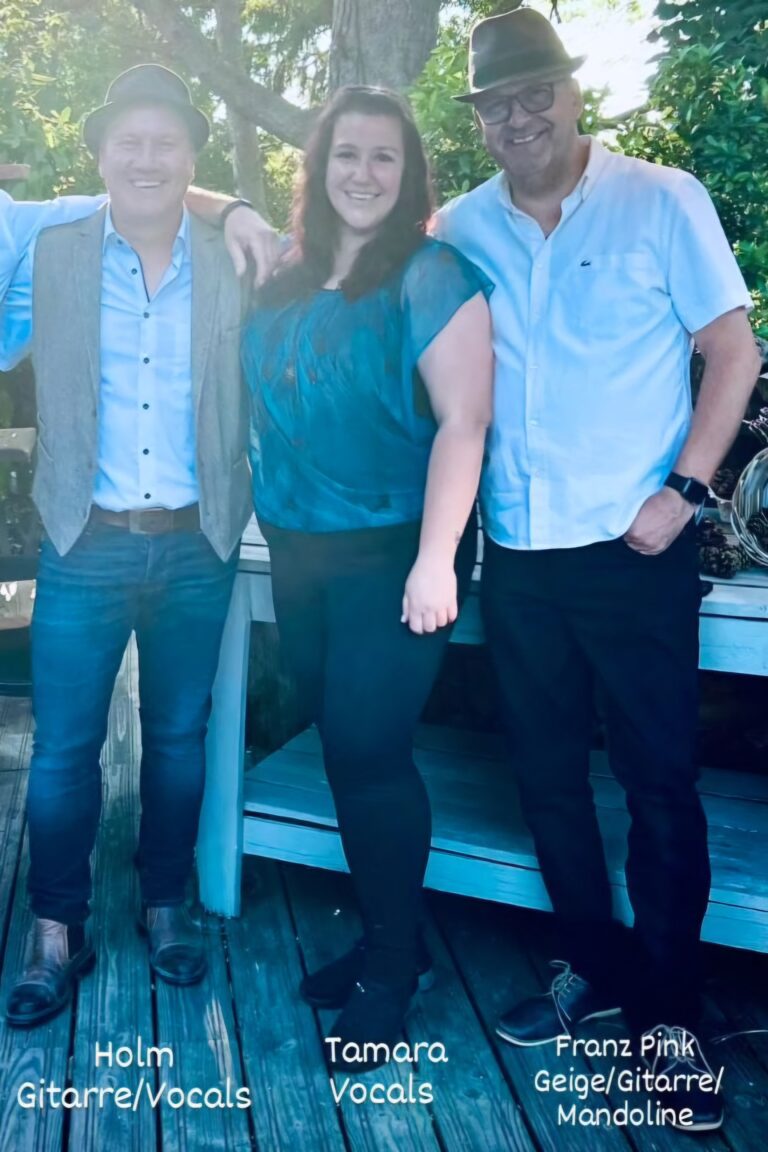Porträt von drei lächelnden Personen auf einer Holzterrasse mit Bäumen im Hintergrund; der Mann links trägt einen Hut, eine Weste, helles Hemd und Jeans; die Frau in der Mitte trägt eine blaue transparente Bluse und schwarze Hosen; der Mann rechts trägt ebenfalls einen Hut, ein weißes Hemd und schwarze Hosen; unter ihren Füßen sind die Namen und Rollen der Personen als Musiker in deutscher Sprache angegeben.