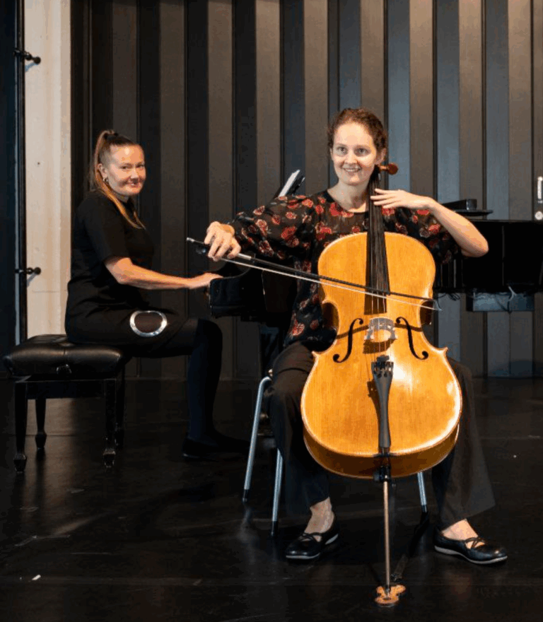 Zwei Frauen auf einer Bühne, eine spielt Cello sitzend, die andere sitzt am Flügel im Hintergrund und lächelt, beide vor einem dunklen vertikal gestreiften Hintergrund.