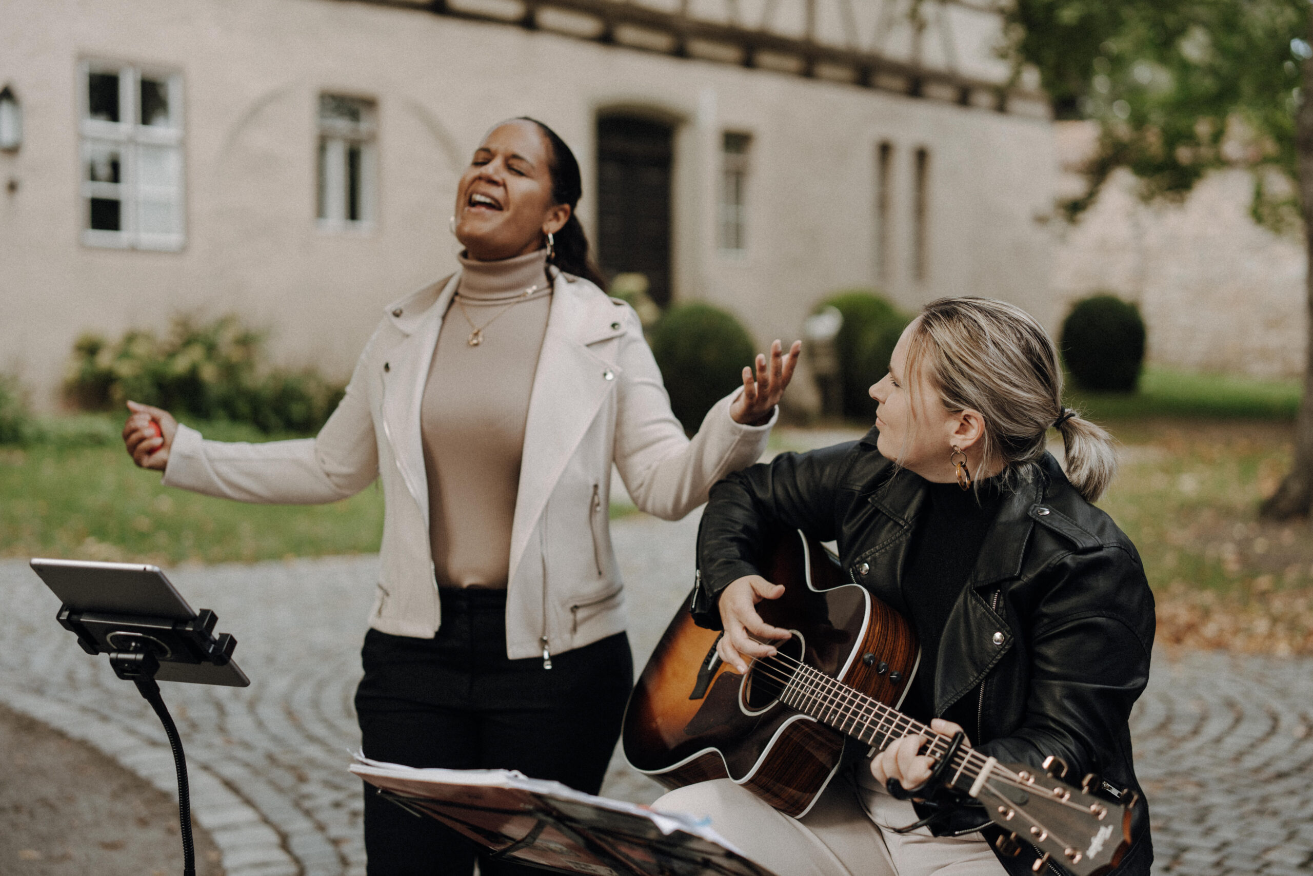 Zwei Frauen musizieren im Freien auf einem gepflasterten Weg vor einem historischen Gebäude, eine singt mit geschlossenen Augen und ausgestreckten Armen, die andere spielt Gitarre und schaut sie an.