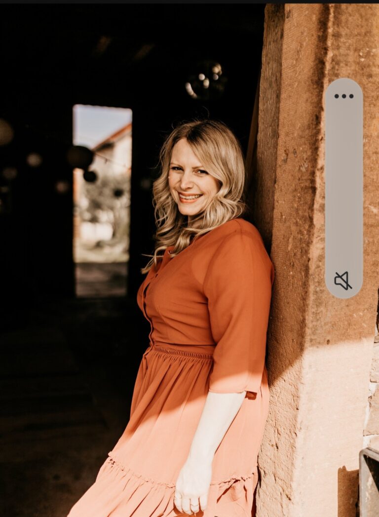 Frau mit langen blonden Haaren in einem orangen Kleid lehnt an einer sonnenbeschienenen Steinmauer und lächelt in die Kamera, im Hintergrund ein dunkler Innenraum mit einem offenen Ausgang nach draußen.