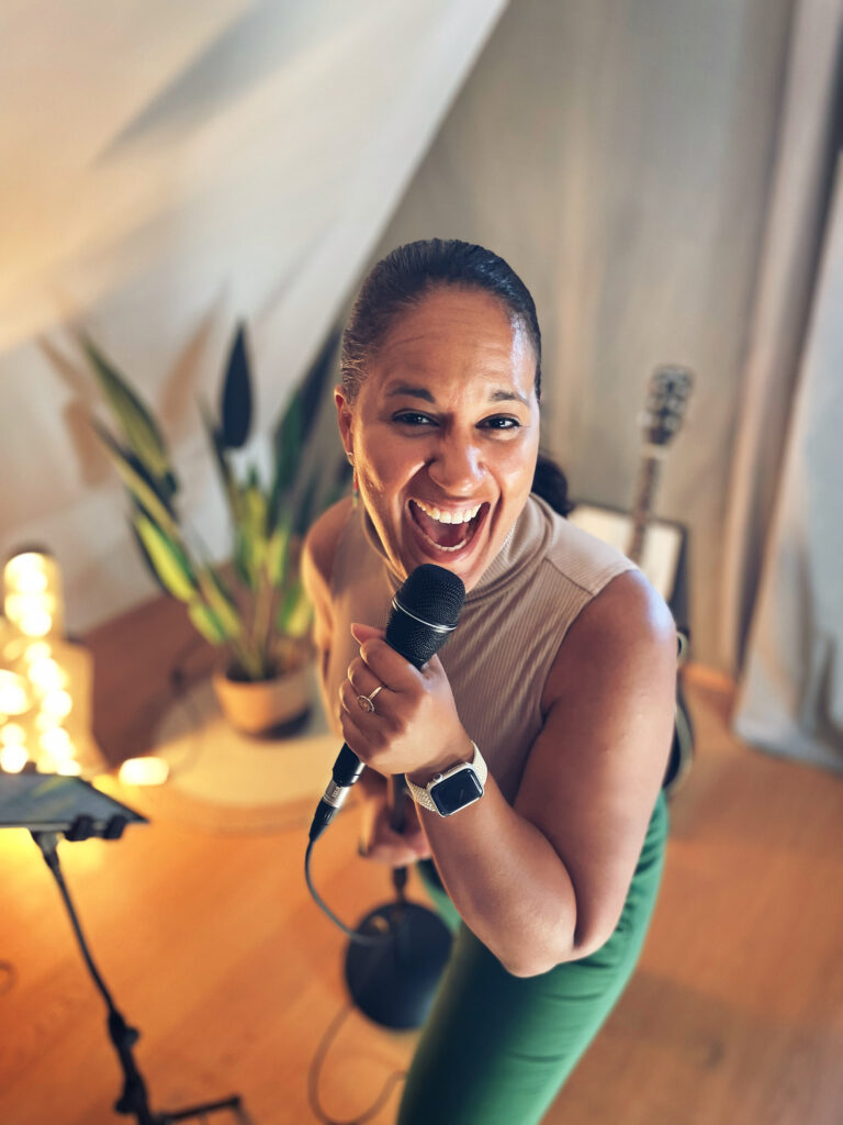 Eine lächelnde Frau hält ein Mikrofon in der Hand und sieht direkt in die Kamera, im Hintergrund sind eine Zimmerpflanze, eine Gitarre und eine helle Lampe zu sehen.