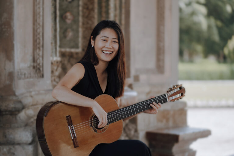 Eine lächelnde Frau mit langen dunklen Haaren, die eine klassische Gitarre spielt, sitzt vor einer verzierten Steinfassade mit unscharfem, grünem Hintergrund.