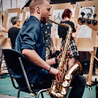 Mann mit dunklem Hemd sitzt auf einem Stuhl und spielt Saxophon in einem Raum, in dem mehrere Personen vor Staffeleien zeichnen.