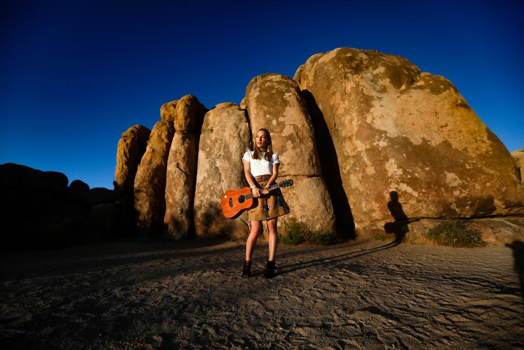 Frau mit weißem T-Shirt und braunem Rock, die vor großen Felsen in der Wüste steht und eine Gitarre hält, bei klarem blauem Himmel und tiefstehender Sonne, die lange Schatten wirft.