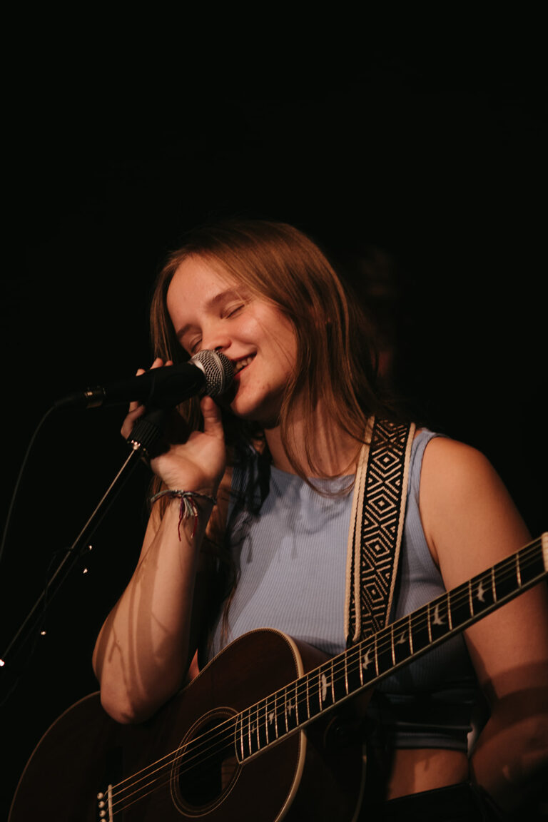 Junge Frau mit langem Haar und blauem Oberteil singt mit geschlossenen Augen in ein Mikrofon und spielt dabei akustische Gitarre vor schwarzem Hintergrund
